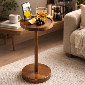 May include: A dark wood side table with a round top and base. A smartphone rests in a slot on the top, next to a glass of amber liquid, a sliced apple, and grapes. The table is a stylish accent piece for a living room.