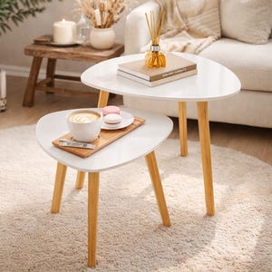 May include: Two white, triangular nesting tables with light wood legs. The tables are arranged in a living room setting, with a cup of coffee and macarons on the lower table. A stack of books and a diffuser sit on the upper table.