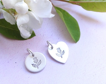May include: Two silver pendants, one round and one heart-shaped, each stamped with a thistle design. The pendants have small loops for hanging. White flowers and green leaves are in the background.