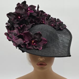 Könnte beinhalten: Ein schwarzer Hut mit breiter Krempe, verziert mit dunkelvioletten Orchideenblüten. Der Hut besteht aus einem gewebten Material und ist für formelle Anlässe konzipiert. Die Blumen sind oben und am Rand angeordnet.