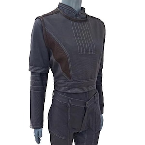 May include: A dark grey two-piece outfit on a mannequin. The top is a cropped, long-sleeved shirt with brown accents and white stitching. The trousers are fitted with brown patches on the knees. The outfit has a futuristic, utilitarian design.