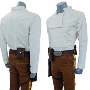 Puede incluir: Un disfraz que presenta una camisa blanca de manga larga con cuello mandarín y pantalones marrones. Los pantalones tienen una raya amarilla en el lateral y se combinan con un cinturón y una funda marrones. El disfraz se exhibe en un maniquí.