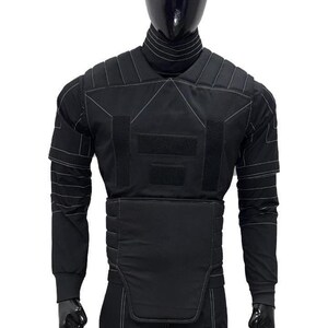 May include: Black tactical vest with white stitching details, displayed on a mannequin. The vest features multiple panels, hook-and-loop closures, and long sleeves. The mannequin is wearing a black turtleneck and pants.