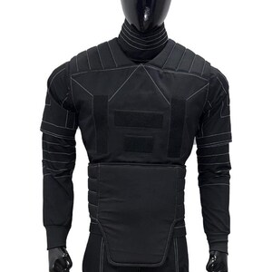 Pu&ograve; includere: Gilet tattico nero con maniche lunghe, esposto su un manichino. Il gilet presenta diversi pannelli con cuciture bianche e chiusure a strappo. Il manichino indossa un dolcevita e guanti neri.