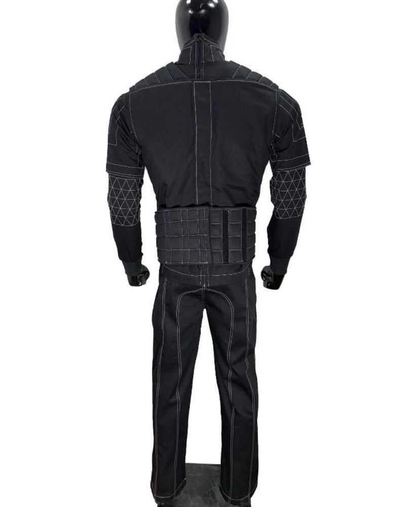 Pu&ograve; includere: Tuta tattica nera con cuciture bianche, esposta su un manichino. La tuta include una giacca con dettagli geometrici sulle maniche, una cintura con pi&ugrave; scomparti e pantaloni abbinati. Il manichino ha una testa nera.