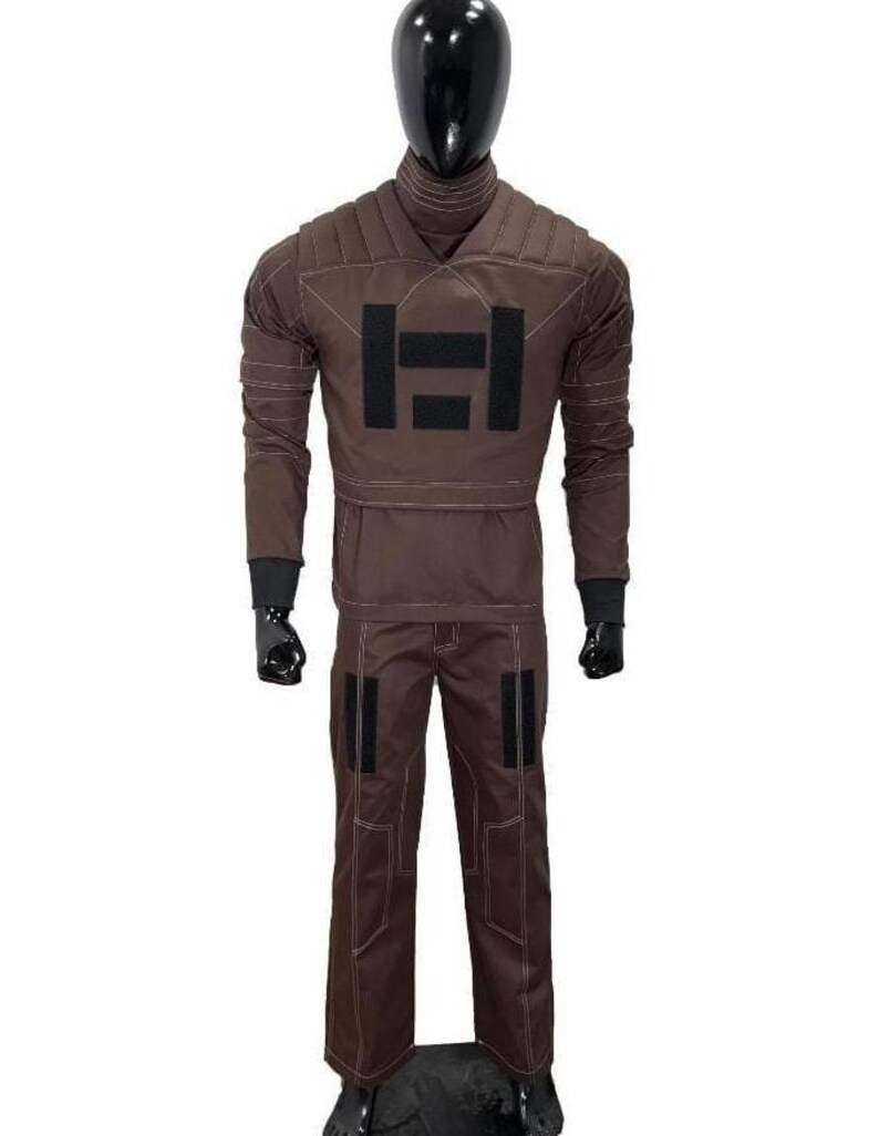 Puede incluir: Mono marr&oacute;n con cuello alto y mangas largas, exhibido en un maniqu&iacute;. El traje presenta un gran dise&ntilde;o rectangular negro en el pecho y parches negros en los pantalones. Las costuras blancas delinean las costuras.