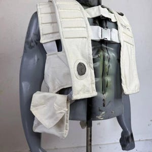 May include: A white vest with shoulder straps, a chest strap, and a small pouch. The vest has a mesh circle on the front and is displayed on a grey mannequin. The vest appears to be made of a durable, possibly protective material.
