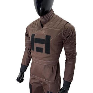 Puede incluir: Mono marr&oacute;n con cuello alto y detalles acolchados en los hombros. El traje presenta detalles en negro y un gran dise&ntilde;o rectangular negro en el pecho. La prenda de manga larga tiene pu&ntilde;os negros y se muestra en un maniqu&iacute;.