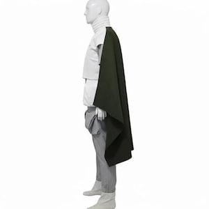 May include: A white high-neck top paired with light grey trousers and a dark olive green cape draped over the shoulder. The ensemble is displayed on a mannequin, showcasing a structured silhouette with clean lines and a layered, monochromatic aesthetic.