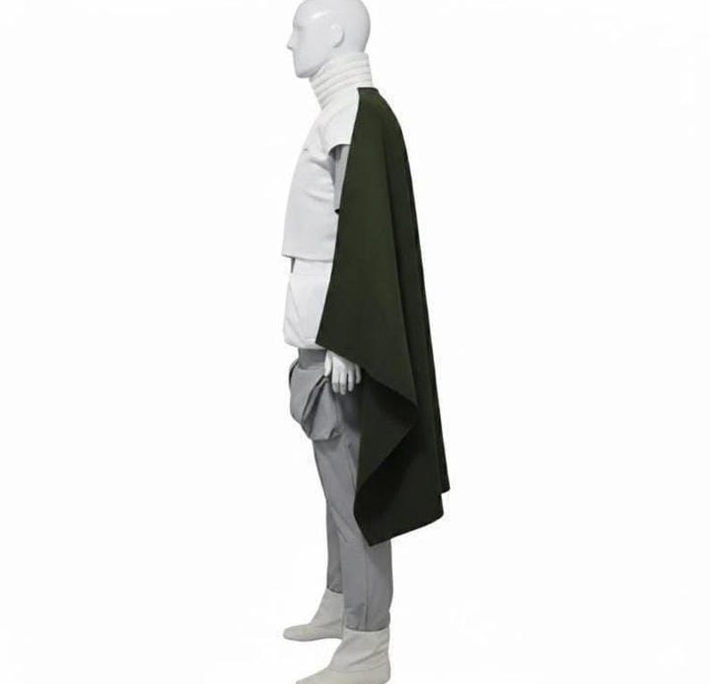 May include: A white high-neck top paired with light grey trousers and a dark olive green cape draped over the shoulder. The ensemble is displayed on a mannequin, showcasing a structured silhouette with clean lines and a layered, monochromatic aesthetic.
