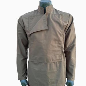 Op de afbeelding: Een bruin-beige shirt met lange mouwen en een uniek asymmetrisch kraagontwerp. Het shirt heeft een hoge kraag en een flap die over de borst kruist. Het kledingstuk wordt op een mannequin getoond, wat de getailleerde pasvorm en stijl benadrukt.