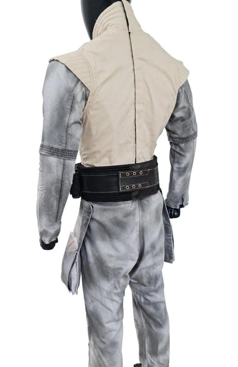 May include: A light gray jumpsuit with a cream-colored vest and a black belt. The jumpsuit has a textured appearance and features pockets on the legs. The vest has a quilted design on the shoulders.