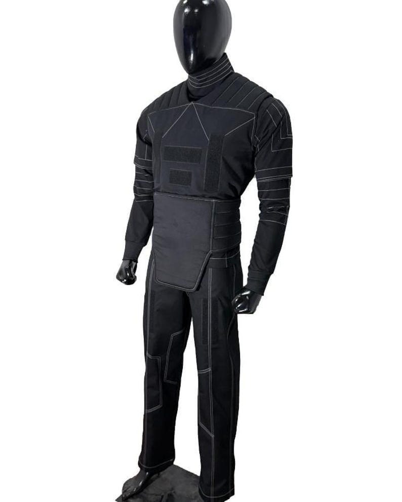 Pu&ograve; includere: Tuta tattica nera con dettagli di cuciture bianche, esposta su un manichino. La tuta comprende un top a maniche lunghe, pantaloni e una protezione per la vita. Il design presenta pannelli rinforzati e un collo alto.