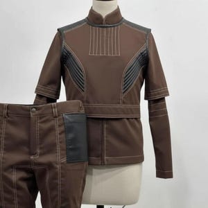 Op de afbeelding: Een bruin tweedelig outfit, bestaande uit een jas en een broek, gepresenteerd op een mannequin. De jas heeft zwarte accenten en witte stiksels. De broek heeft zwarte patches en witte stiksels.