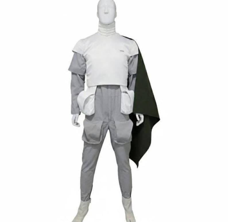 May include: A white and light grey jumpsuit featuring a high collar, short sleeves, and a white chest plate. The garment includes four large utility pockets on the hips and thighs, paired with a dark olive green cape draped over the left shoulder.