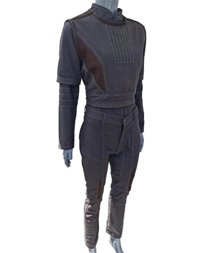 May include: A dark grey two-piece outfit on a mannequin. The top is a cropped, long-sleeved shirt with brown accents and white stitching. The trousers are fitted with brown patches on the knees. The outfit has a futuristic, utilitarian design.