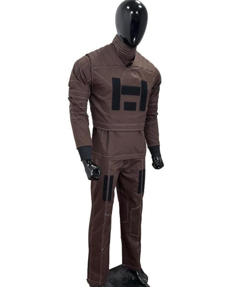 Puede incluir: Mono marr&oacute;n con detalles negros y costuras blancas. La parte superior de manga larga presenta un cuello alto y un gran dise&ntilde;o rectangular negro en el pecho. Los pantalones tienen parches rectangulares negros en las piernas. El maniqu&iacute; tiene una cabeza negra y guantes negros.