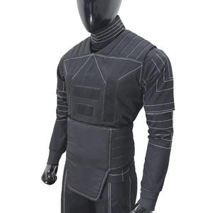 Pu&ograve; includere: Set di gilet e pantaloni tattici neri esposto su un manichino. Il gilet presenta cuciture bianche, pannelli multipli e chiusure a strappo. La camicia a maniche lunghe e i pantaloni sono anch'essi neri, con dettagli di cucitura bianchi.