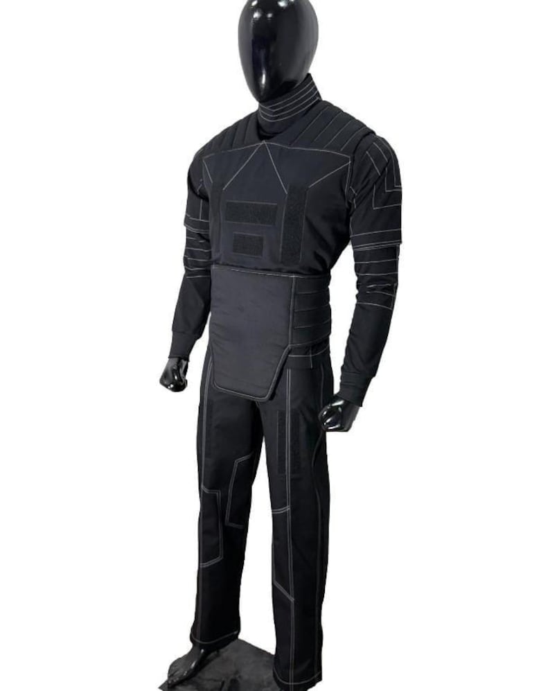 Pu&ograve; includere: Tuta tattica nera con dettagli di cuciture bianche. La tuta presenta un collo alto, maniche lunghe e un design segmentato sul petto e sulla vita. I pantaloni hanno un motivo cucito simile. Esposto su un manichino.