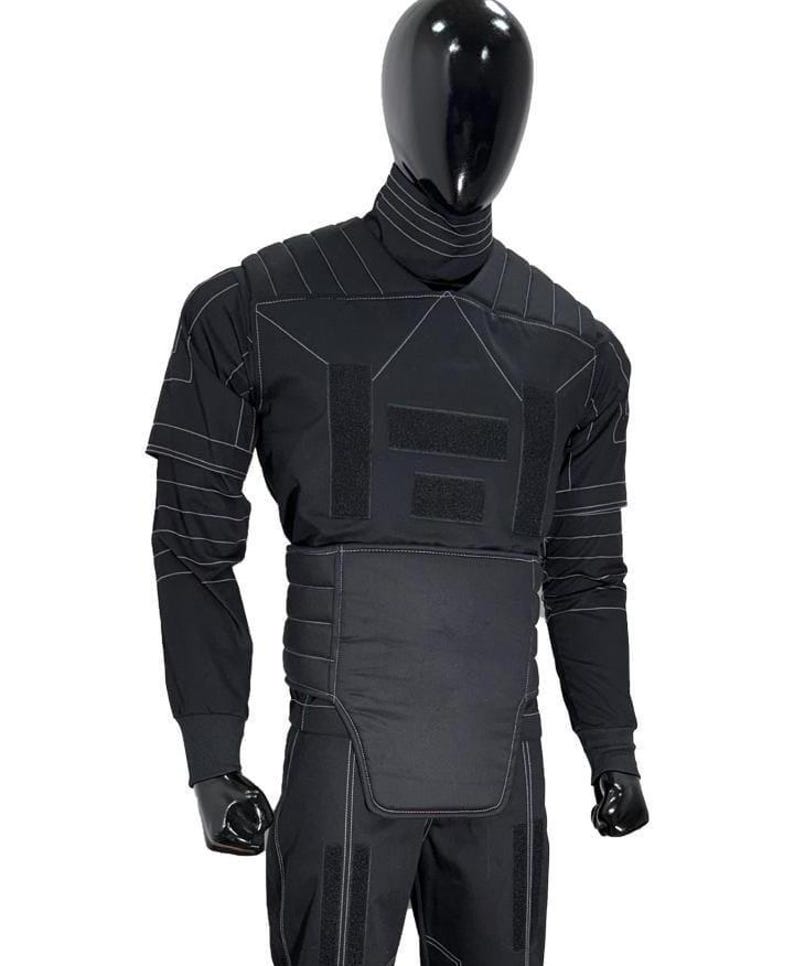 May include: Black tactical suit displayed on a mannequin. The suit features a high collar, long sleeves, and reinforced panels with white stitching. The design includes multiple hook-and-loop fastener patches and a padded lower torso section.