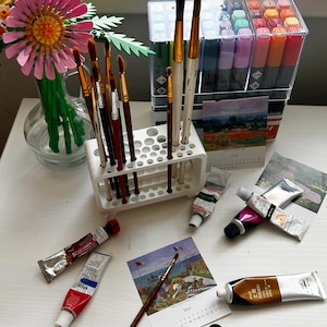 Può includere: Una collezione di materiali artistici, tra cui pennelli, tubetti di vernice e pennarelli, disposti su una superficie bianca. Sono presenti anche un portapennelli bianco e un calendario con un dipinto. Un vaso di fiori colorati è sullo sfondo.