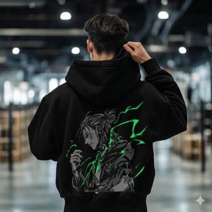 May include: Black hoodie featuring a graphic of an anime character with green lightning accents on the back. The character is depicted in a side profile with detailed facial features and a bun hairstyle. The hoodie has a drawstring hood and ribbed cuffs.