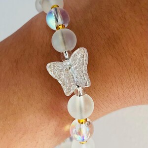 May include: A clear beaded bracelet featuring a butterfly charm. The bracelet has iridescent and frosted white beads with gold-colored accents. The butterfly charm is clear and has a detailed design.