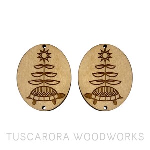May include: Two wooden round ornaments with a laser-cut design of a turtle with a plant growing from its back. The plant has a star at the top.