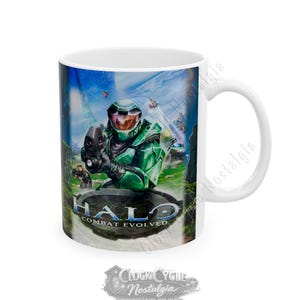 May include: A white ceramic mug featuring a colorful graphic of a green armored soldier holding a weapon from the Halo Combat Evolved video game. The design includes the Halo Combat Evolved logo at the bottom, set against a blue sky background.
