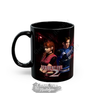 May include: A black ceramic mug featuring a graphic print of the video game Resident Evil 2. The design displays two characters, one in a pink vest and one in a blue uniform, with the Resident Evil 2 logo printed in white and red text.