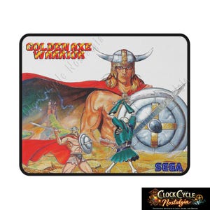 May include: Rectangular mousepad featuring the Golden Axe Warrior video game cover art. The design includes a muscular warrior with a horned helmet, a shield, and two other figures in combat. The SEGA logo is visible. The mousepad has a black border.