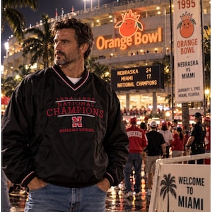 May include: A black long-sleeve pullover jacket featuring a V-neck collar with red and white trim. The chest displays the text 1994-1995 NATIONAL CHAMPIONS, a red N logo, and the text NEBRASKA HUSKERS in red lettering on a black background.