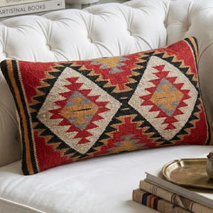 May include: Rectangular decorative pillow with a geometric pattern in red, black, beige, and yellow. The pillow is placed on a white tufted sofa. Books and a small bird figurine are also visible.