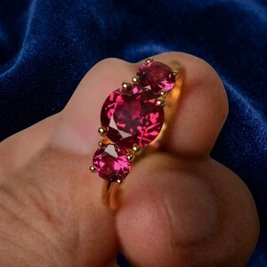 May include: A gold ring featuring three round-cut, vibrant pinkish-red gemstones set in a row. The center stone is the largest, flanked by two smaller stones of the same color, all secured by gold prongs on a polished gold band.