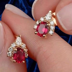 May include: A pair of gold earrings, each featuring a central oval-cut red gemstone surrounded by a cluster of small, clear, round-cut diamonds and a single pear-cut diamond at the top, set in a polished gold metal frame.