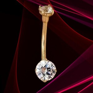 May include: A gold-coloured belly button ring featuring two clear, round-cut gemstones. The top gem is smaller and set in a prong setting, while the bottom gem is larger. The curved bar connects the two gems.