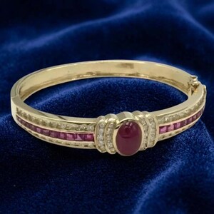 May include: A gold hinged bangle bracelet featuring a central oval cabochon ruby surrounded by small round diamonds. The band is accented with a channel-set row of square-cut rubies and additional small diamonds along the sides of the metal frame.