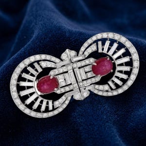 Può includere: Una spilla color argento con due pietre rosse ovali a cabochon al centro. Il pezzo è decorato con numerose piccole pietre chiare a taglio rotondo e pietre rettangolari a taglio baguette disposte in un motivo geometrico simmetrico.