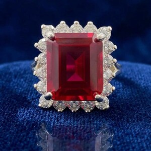 Peut inclure: Une bague ornée d'une grande pierre précieuse rouge foncé de taille émeraude rectangulaire au centre, entourée d'un halo de petites pierres blanches rondes et claires serties sur une bande métallique. Le design met en valeur une pierre rouge.
