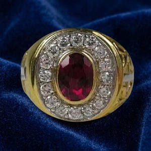 May include: A gold ring featuring a central oval-cut deep red gemstone surrounded by a halo of small, round, clear white stones. The band is crafted from polished yellow gold, providing a contrast to the vibrant red center and the bright halo.