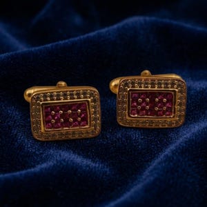 May include: A pair of rectangular gold-toned cufflinks featuring a central grid of deep red square-cut stones surrounded by a border of small, clear, sparkling crystals set within a metallic frame with a classic hinged toggle backing.