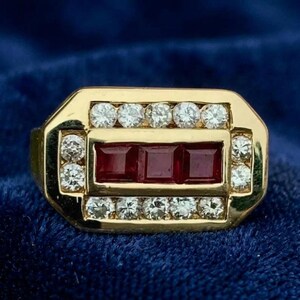 May include: A gold ring featuring a rectangular center set with three square-cut red rubies. The rubies are surrounded by a border of small, round, clear white diamonds set into the gold frame, creating a geometric, multi-stone design.