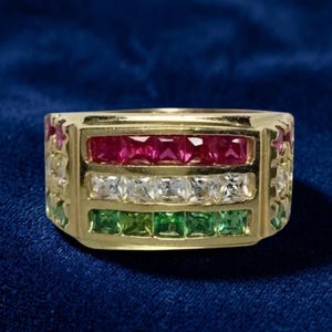 May include: A gold band ring featuring three horizontal rows of square-cut gemstones. The top row contains red stones, the middle row contains clear white stones, and the bottom row contains green stones, with additional stones set along the sides.