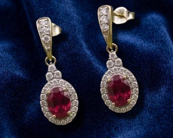 May include: A pair of gold drop earrings featuring oval-cut red gemstones surrounded by a halo of small white stones. The top portion of each earring is tapered and set with three additional white stones, connecting to a post-style back closure.