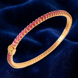May include: A gold-toned bangle bracelet featuring a continuous row of small, round, vibrant pink stones set along the outer edge. The bracelet includes a secure hinged clasp mechanism on one side, maintaining a circular shape throughout.