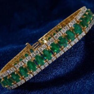 May include: A gold bracelet featuring a central row of oval-cut green emeralds flanked by two parallel rows of small, clear, round-cut white stones. The piece includes a secure metal clasp mechanism visible at the end of the band structure.