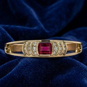 Op de afbeelding: Een gouden ring met een centrale rechthoekige rode edelsteen, geflankeerd door drie kleine ronde witte diamanten aan elke kant. De ring heeft een gladde, gepolijste afwerking en een breed, opengewerkt ontwerp dat de stenen horizontaal omlijst.