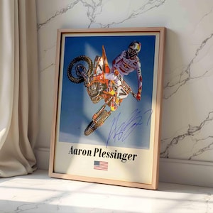 May include: A framed poster featuring a motocross rider on an orange motorcycle mid-air against a blue sky. The print includes a blue ink signature, the name Aaron Plessinger in black text, and a small United States flag icon at the bottom.