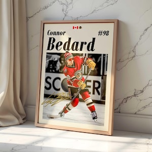 May include: Framed print featuring hockey player Connor Bedard in a red and black uniform, with the number 98. The print includes the Canadian flag and the word "SCORES". The frame is a light wood color.