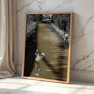 Puede incluir: Fotografía en blanco y negro enmarcada de un golfista en pleno swing en un campo de golf. La imagen está en un marco de madera y se apoya en una pared de mármol blanco. El golfista lleva gorra, camisa y pantalón blancos.